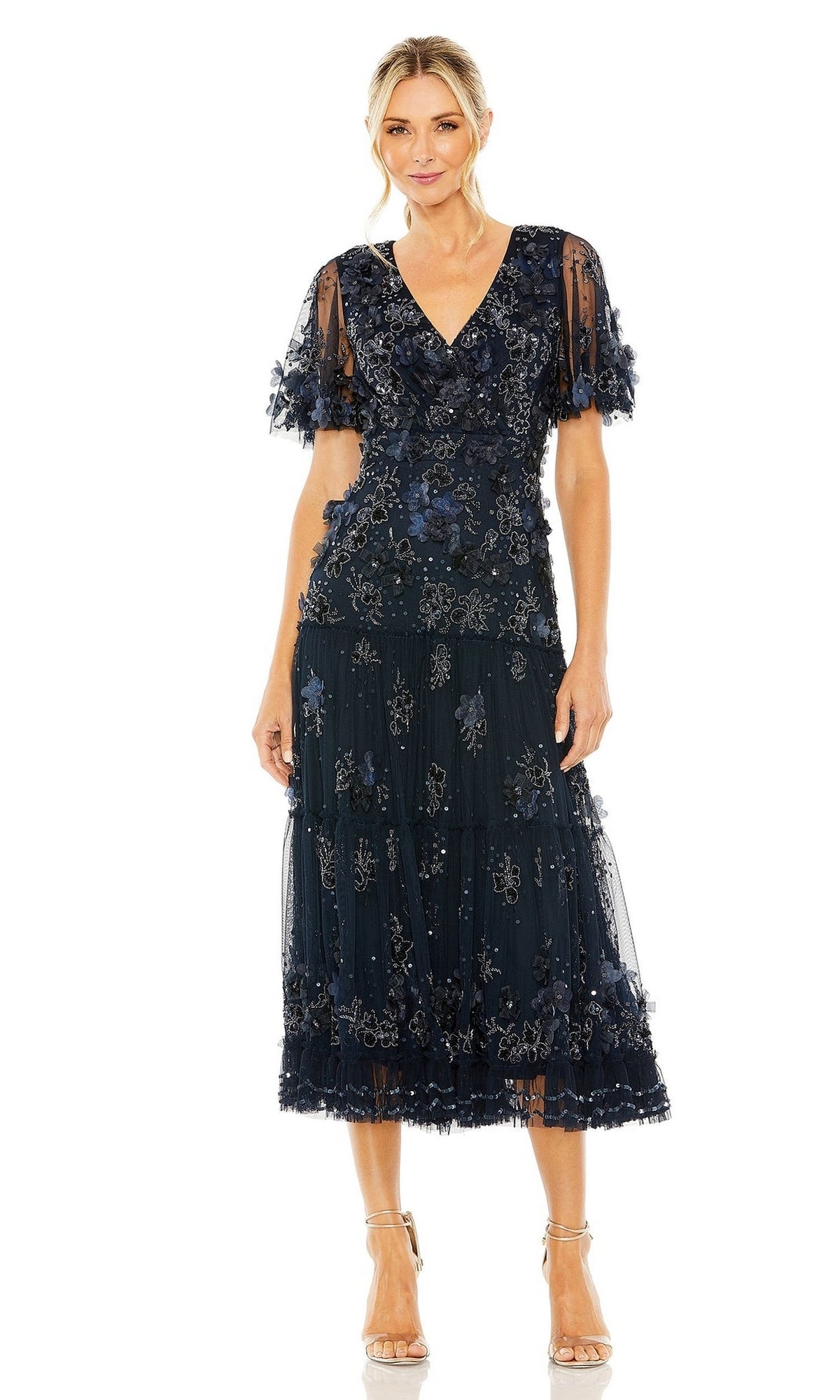 A woman wears the Mac Duggal 9207 A-Line Wedding Guest Dress in navy blue with floral embellishments and short sheer sleeves, styled with neutral-heeled sandals against a white background.